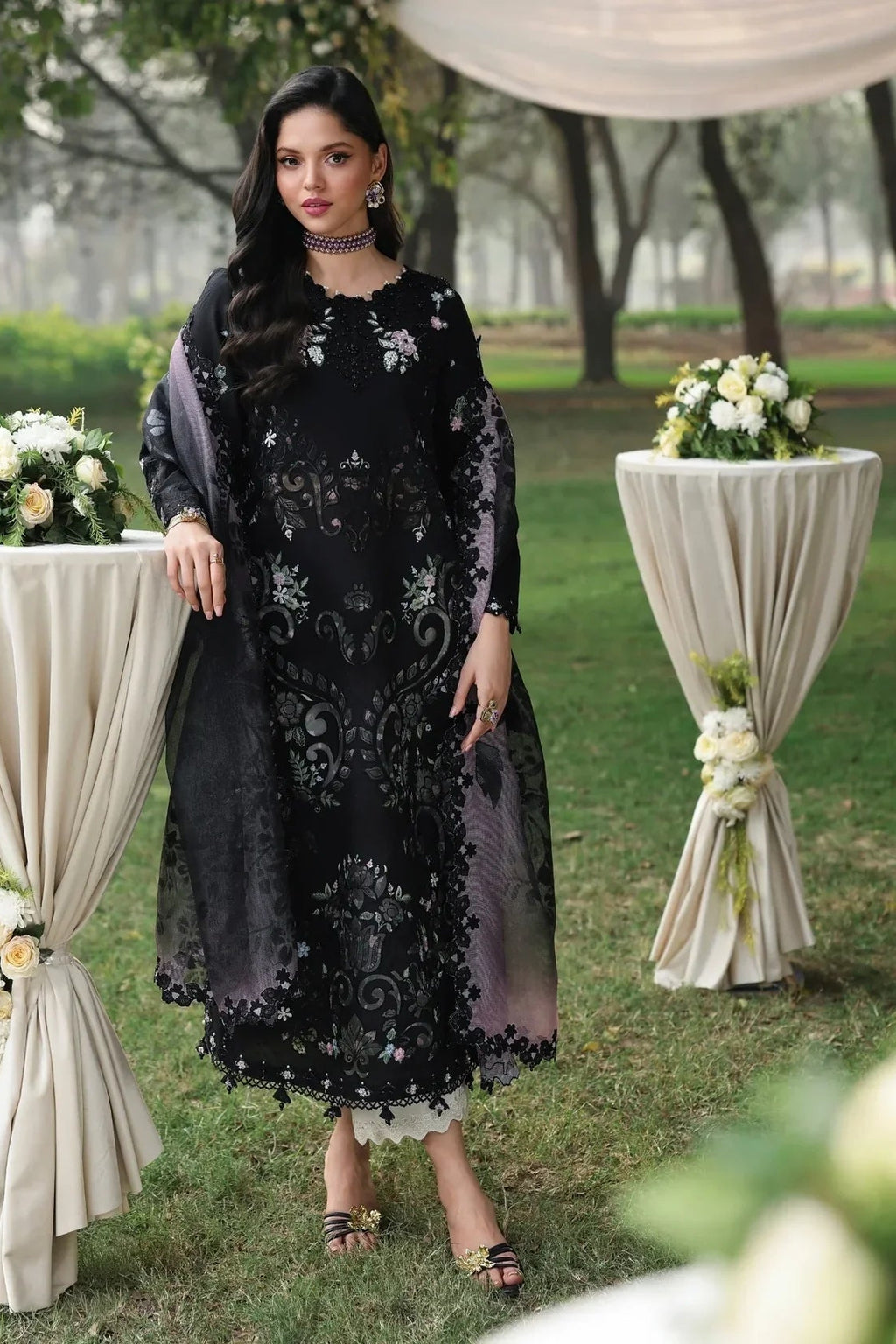 Woman in a black embroidered dress standing outdoors with floral decorations.