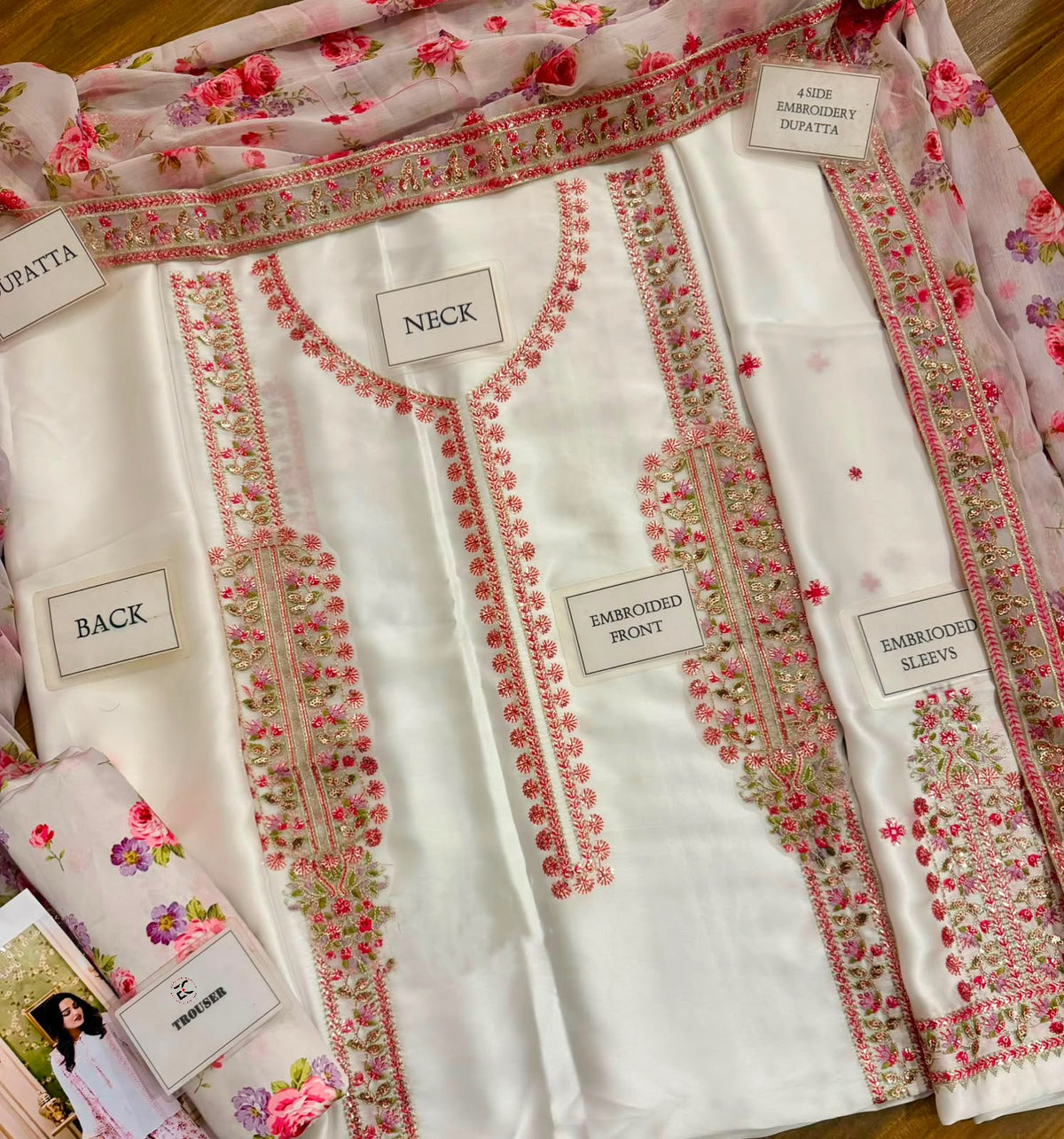 A folded white traditional kurta set with pink floral embroidery and matching sheer dupatta, laid flat on a wooden surface. Multiple fabric labels are visible, including NECK, BACK, BAMBINED FRONY, BAMBINED SLOZY, and LACE BAMBINED DUPatta.