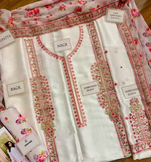 A folded white traditional kurta set with pink floral embroidery and matching sheer dupatta, laid flat on a wooden surface. Multiple fabric labels are visible, including NECK, BACK, BAMBINED FRONY, BAMBINED SLOZY, and LACE BAMBINED DUPatta.