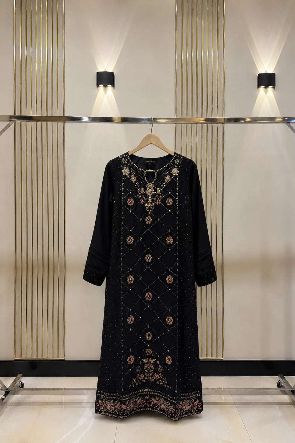 A black long-sleeved tunic dress with intricate gold and brown embroidery, hanging on a wooden hanger on a clothing rack against a modern wall with vertical gold stripes and wall lights.