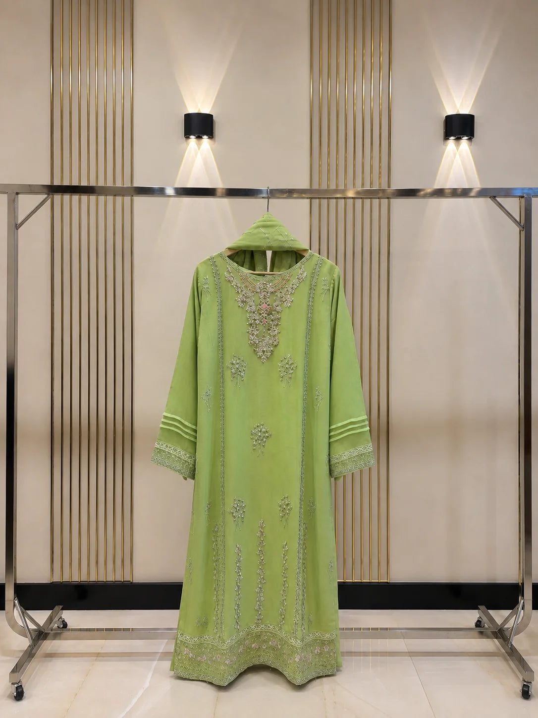 A light green traditional long kurta with intricate silver embroidery, hanging on a metal clothing rack against a beige slatted wall with wall-mounted lights.