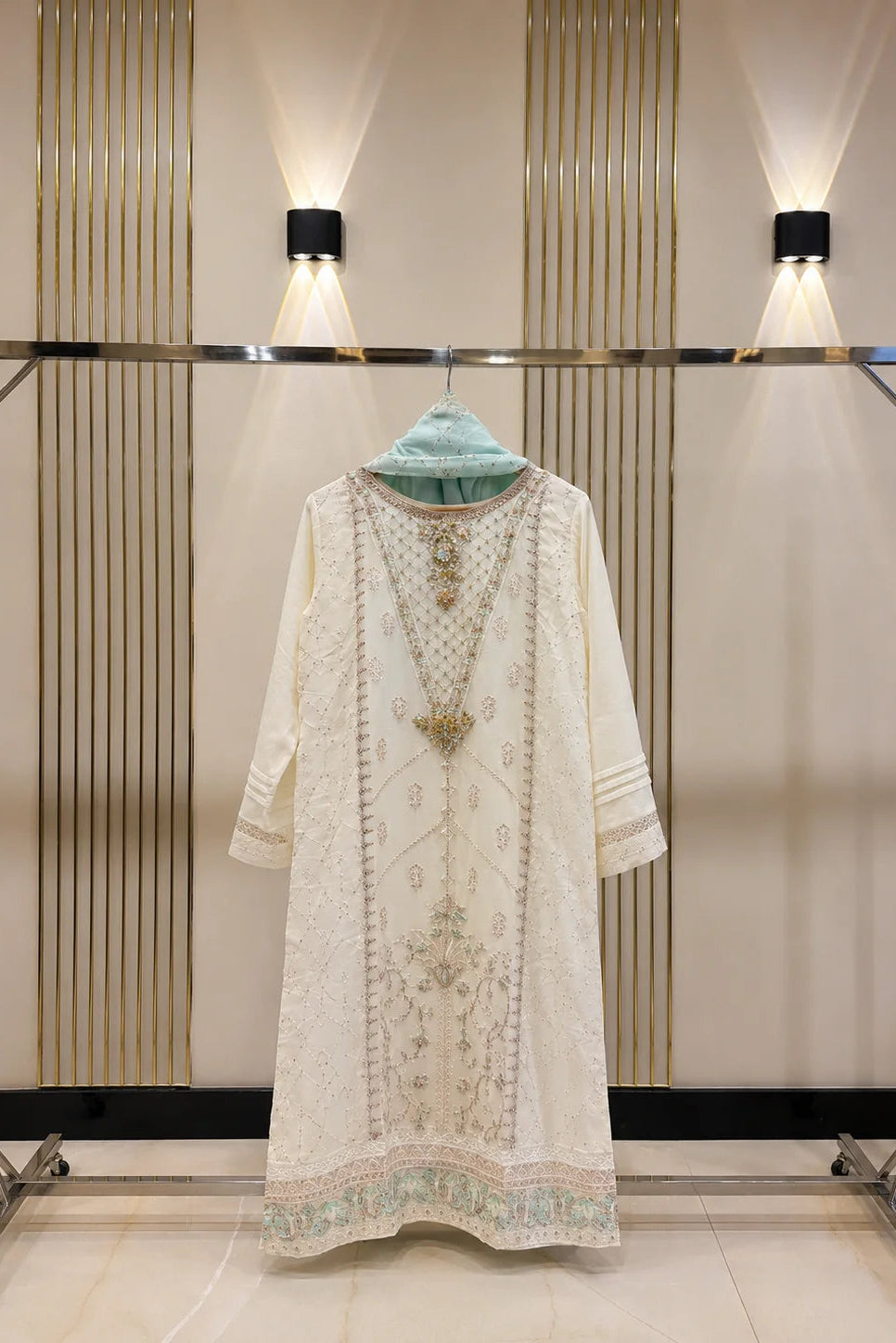 An off-white embroidered kurta with gold and mint green detailing, displayed on a clothing rack with a mint green headscarf.