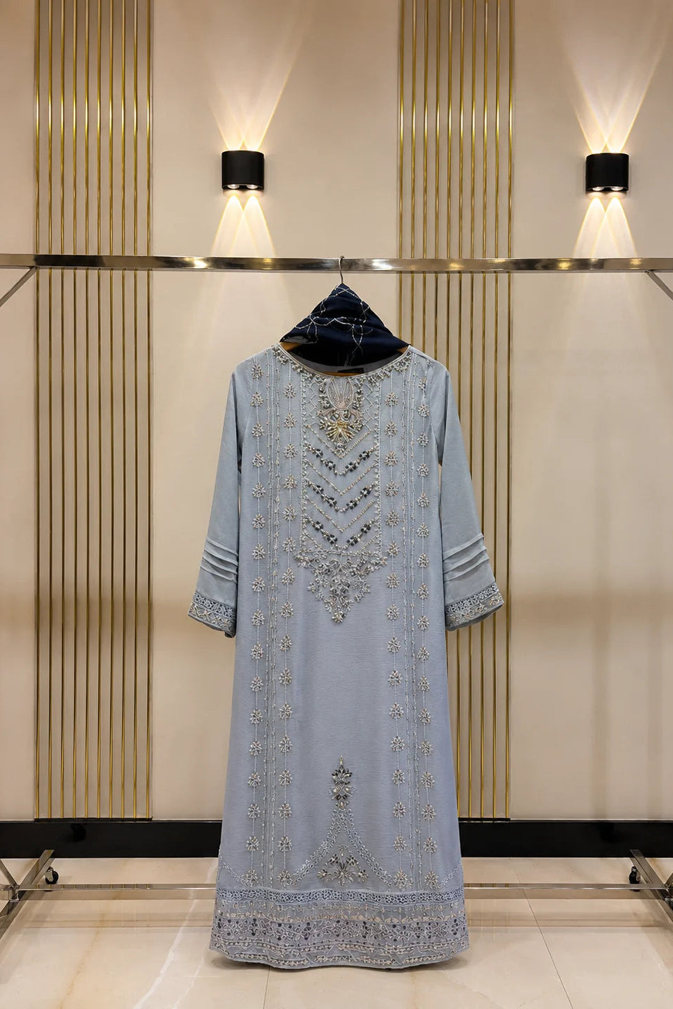 A light blue traditional long dress with intricate silver and gold embroidery, displayed on a black hanger on a clothing rack against a beige wall with vertical gold stripes and wall lights.
