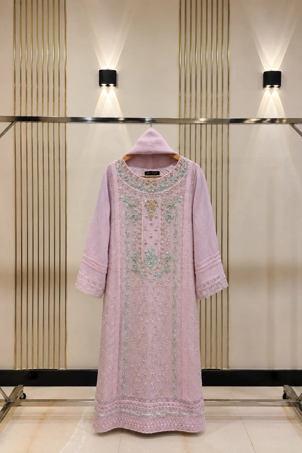 A light pink traditional long tunic dress with intricate green and gold embroidery, displayed on a clothing rack against a beige wall with vertical wood slats and wall-mounted lights.