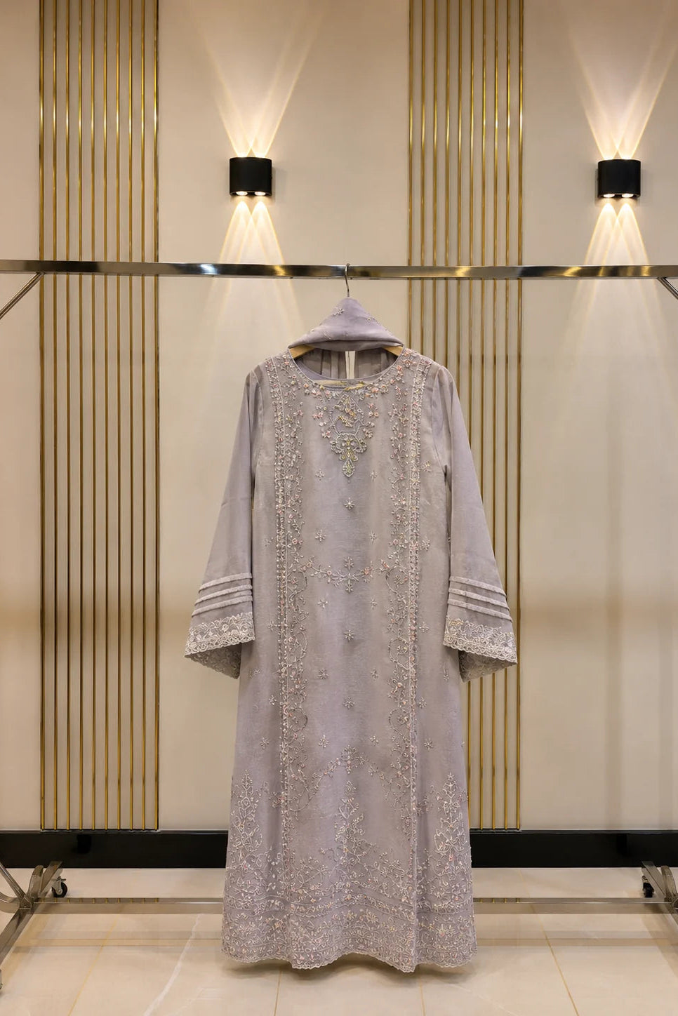 A light lavender-gray traditional long dress with intricate silver embroidery, displayed on a clothing rack against a beige wall with vertical gold slats and wall lights.