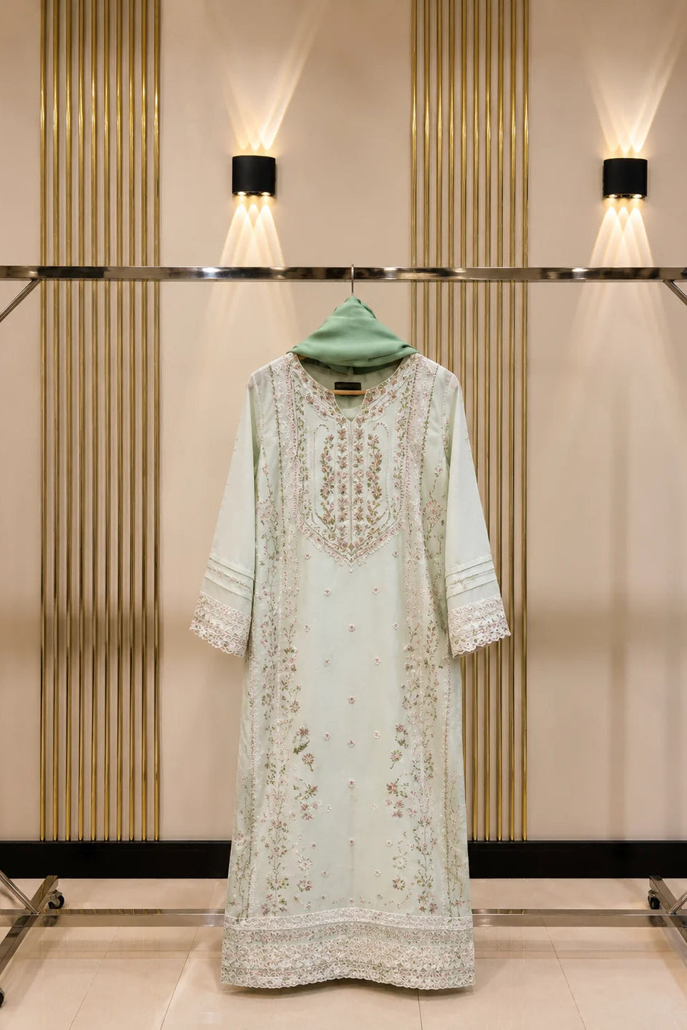 A light sage green traditional long kurta with intricate gold embroidery, displayed on a metal clothing rack against a beige wall with vertical gold slats and wall-mounted lights.
