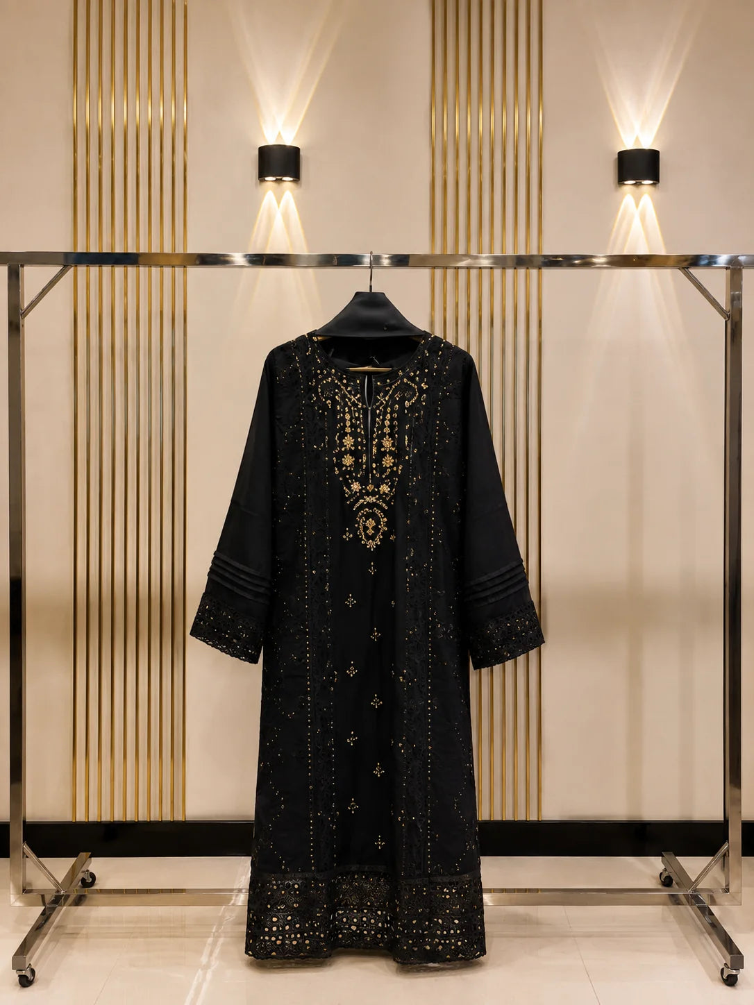 A black long-sleeved tunic or kaftan with intricate gold embroidery and beadwork, displayed on a black hanger on a clothing rack against a beige wall with vertical gold slats and wall lights.