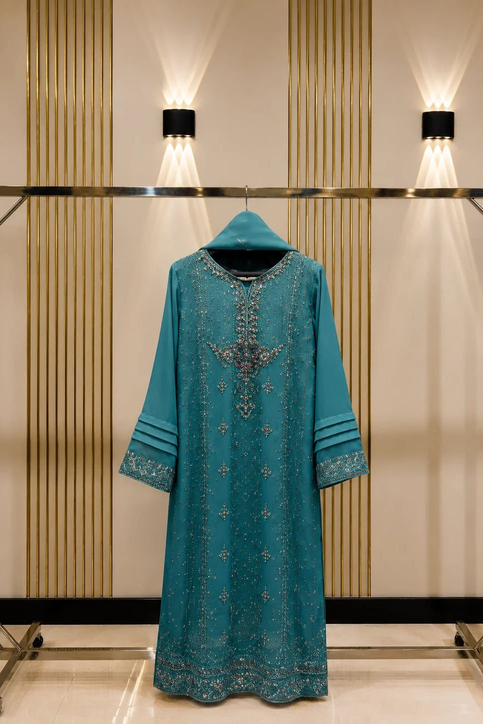 A teal blue long kurta with intricate silver embroidery and a hood, displayed on a clothing rack against a beige wall with vertical gold stripes and wall-mounted lights.