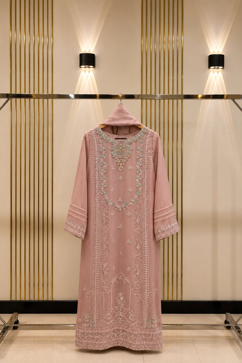 A dusty pink embroidered kurta with a matching hood, hanging on a clothing rack against a beige wall with vertical gold stripes and two wall-mounted lights.