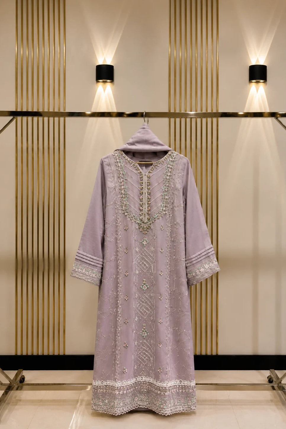 A lavender purple traditional long tunic dress with intricate silver and white embroidery, hanging on a gold metal clothing rack against a beige wall with vertical slats and wall-mounted lights.