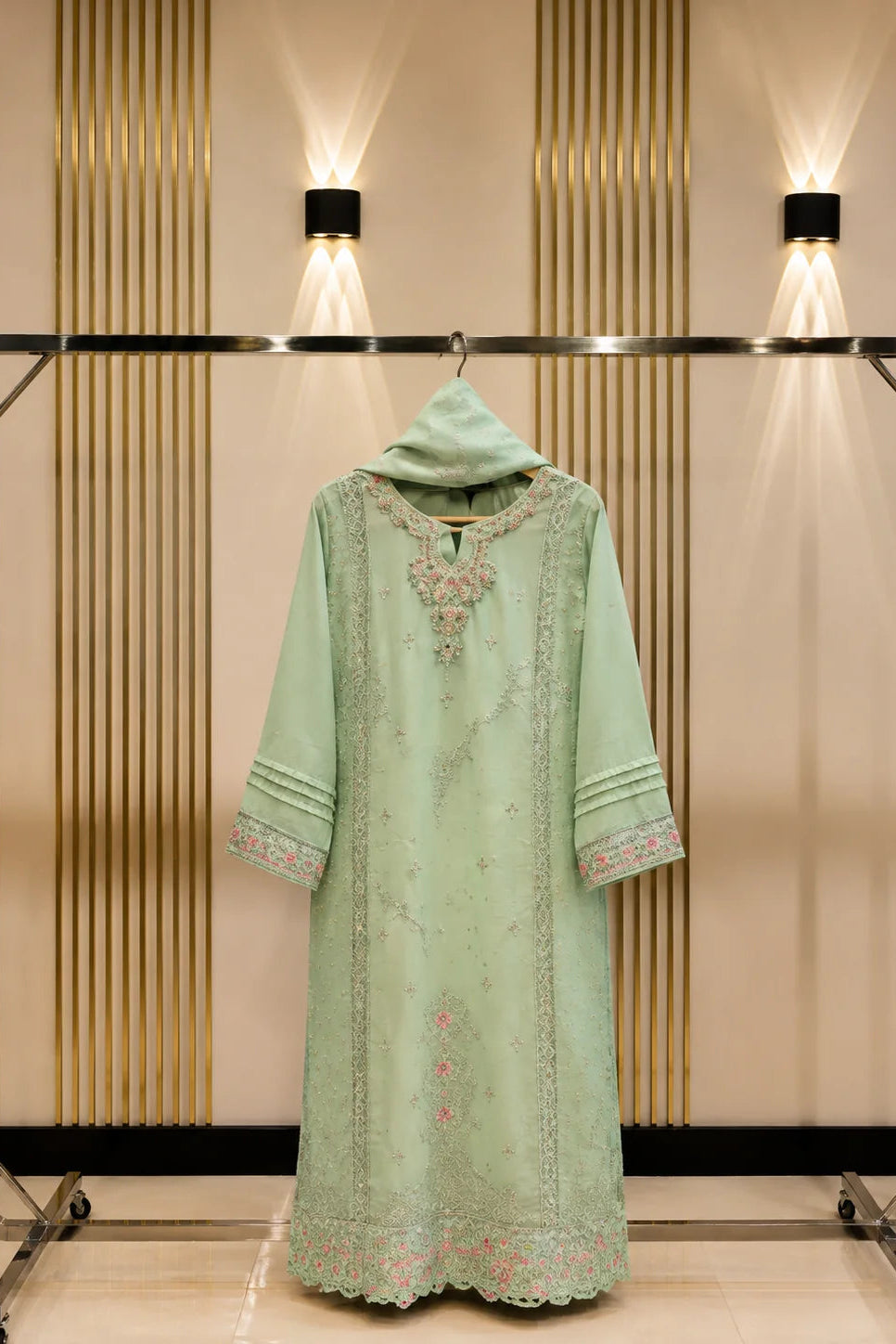 A mint green traditional long kurta with floral embroidery and a matching hood, displayed on a clothing rack against a beige wall with vertical gold panels and wall lights.