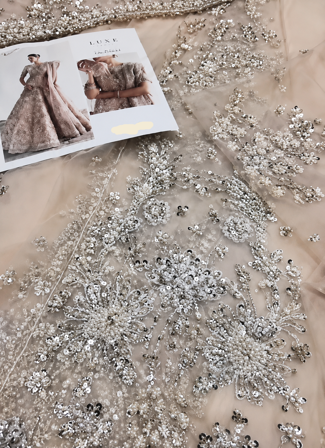 Close-up of a beige tulle garment or fabric with extensive silver beadwork and sequin embroidery, with a printed fashion magazine page placed on top.