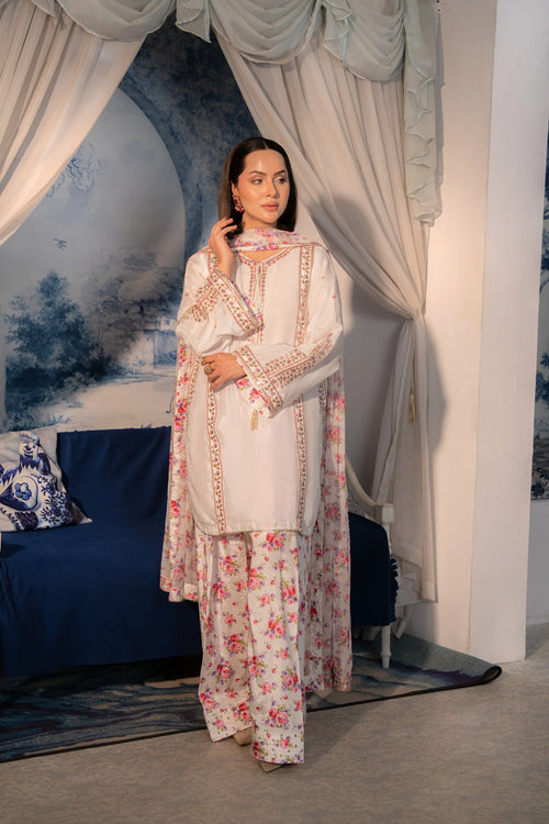 A woman models a traditional South Asian outfit consisting of a cream-colored kurta with gold embroidery, a matching floral dupatta, and matching printed trousers, set against a backdrop of draped curtains and a blue sofa.