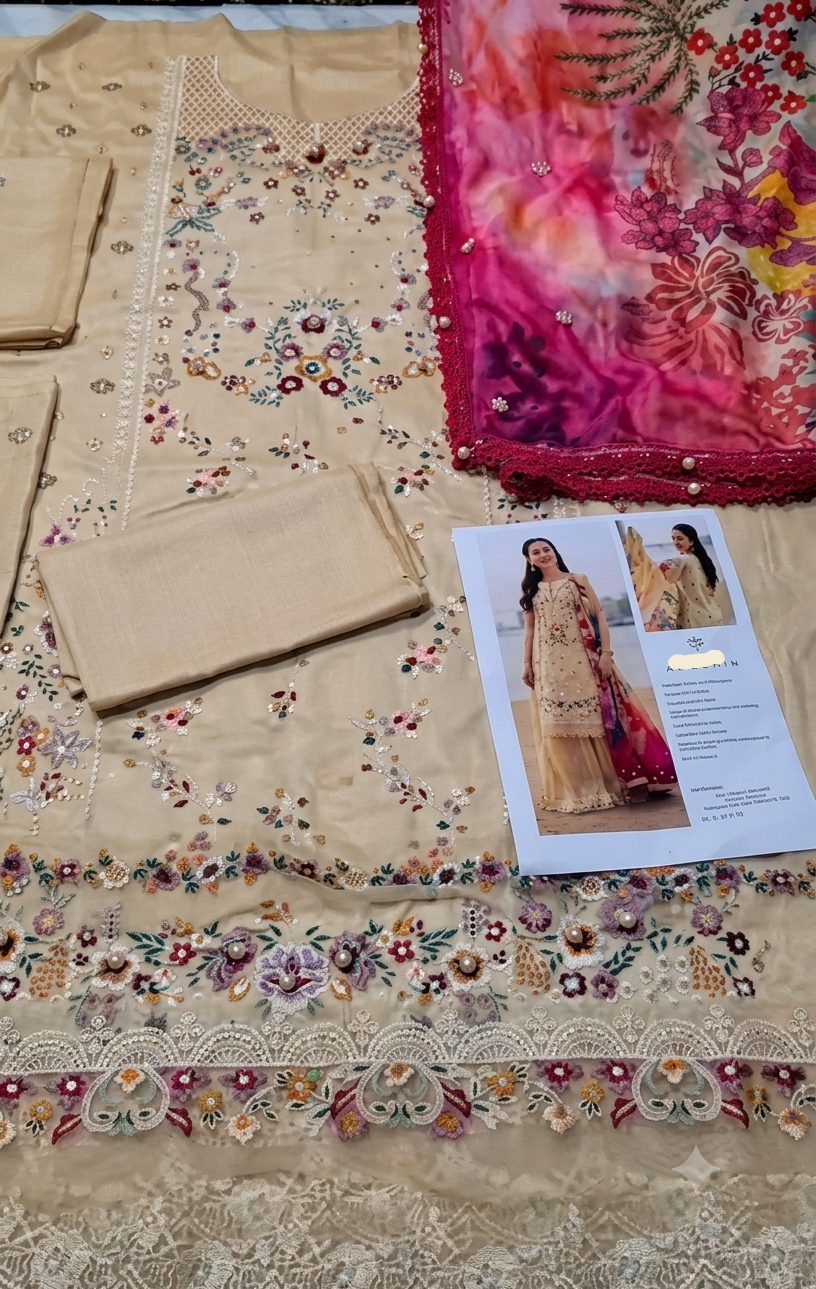 A beige embroidered traditional outfit set laid flat, including a kurta with floral embroidery, matching fabric pieces, and a bright pink floral dupatta. A printed garment card is placed on top of the outfit.