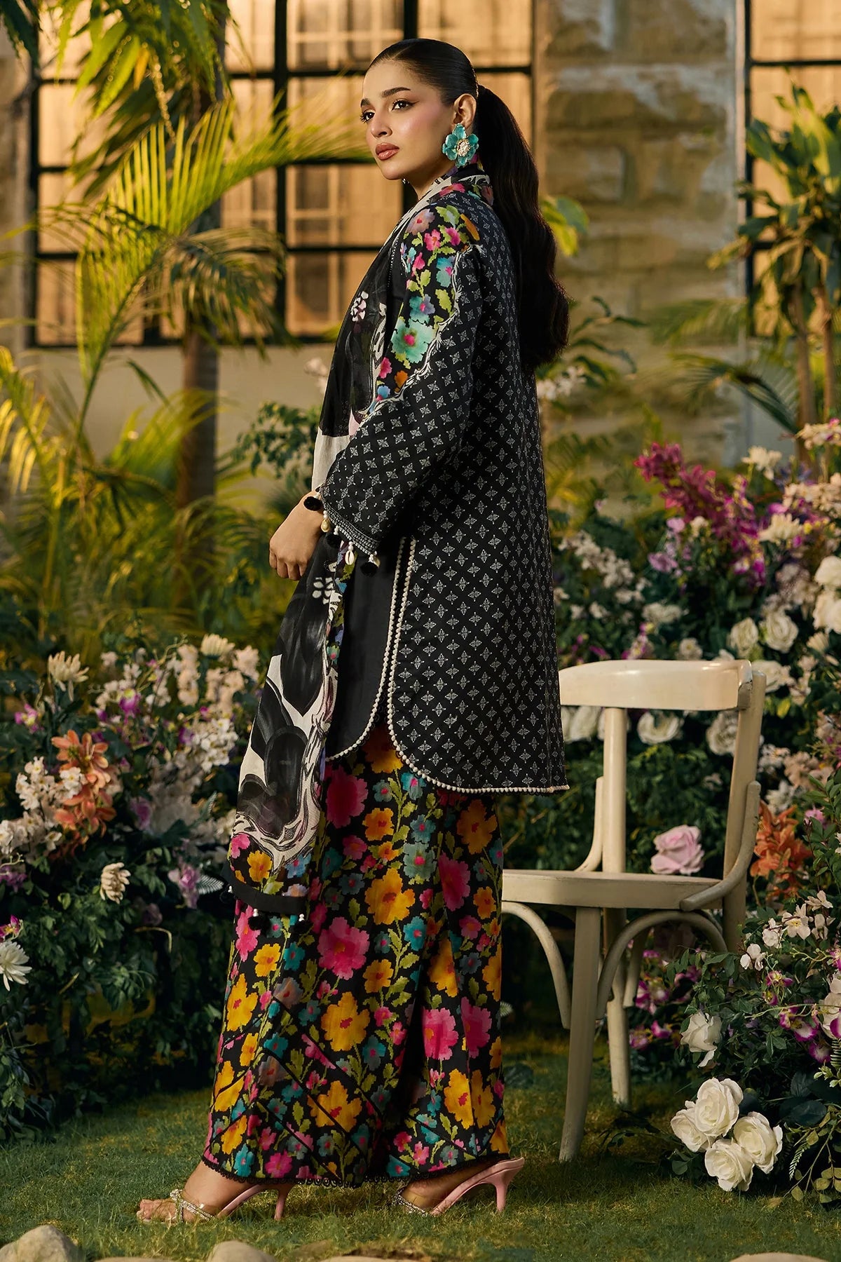 A woman models a traditional South Asian outfit in a garden setting. The outfit consists of a black patterned jacket with colorful floral embroidery on the sleeves, paired with matching wide-leg trousers featuring a vibrant multicolor floral print.