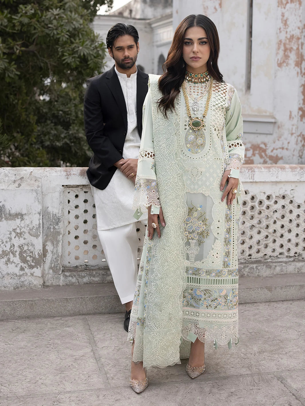 Man and woman in formal attire standing outdoors with a decorative wall and trees in the background.