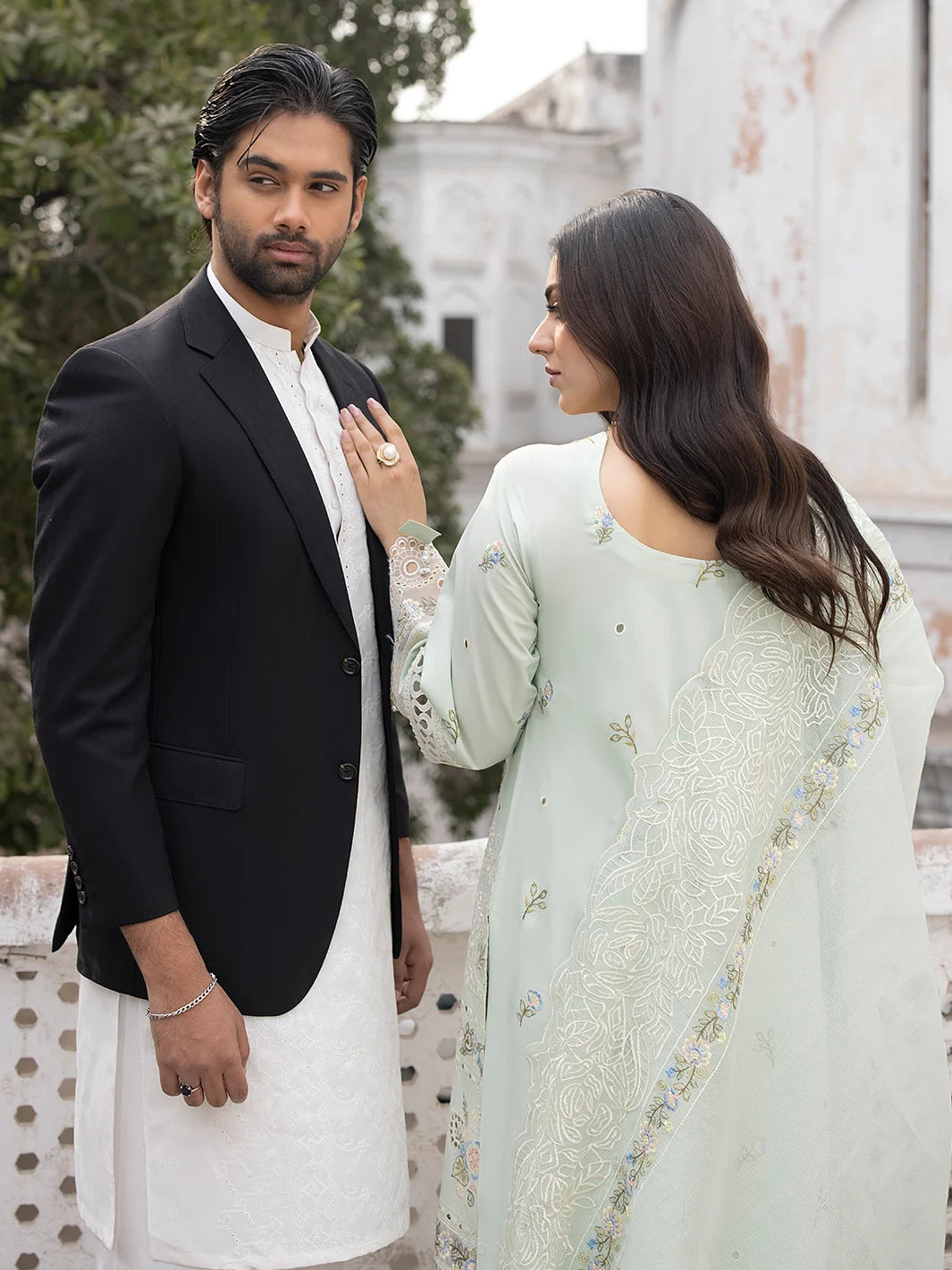 Man in black suit and white pants, woman in light green traditional outfit with floral patterns, standing outdoors.