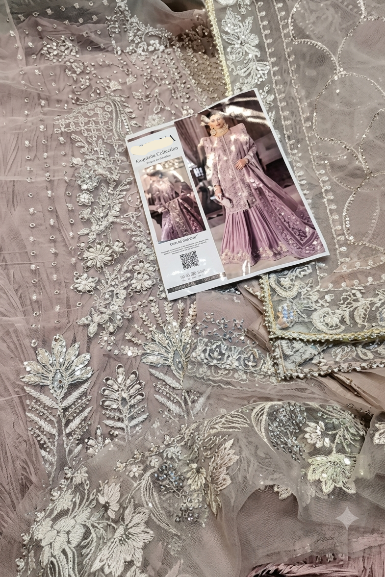 An ornate light pink or dusty rose embroidered traditional garment laid flat, featuring intricate silver beadwork, sequins, and lace. A Soraya brand card is placed on top of the fabric.